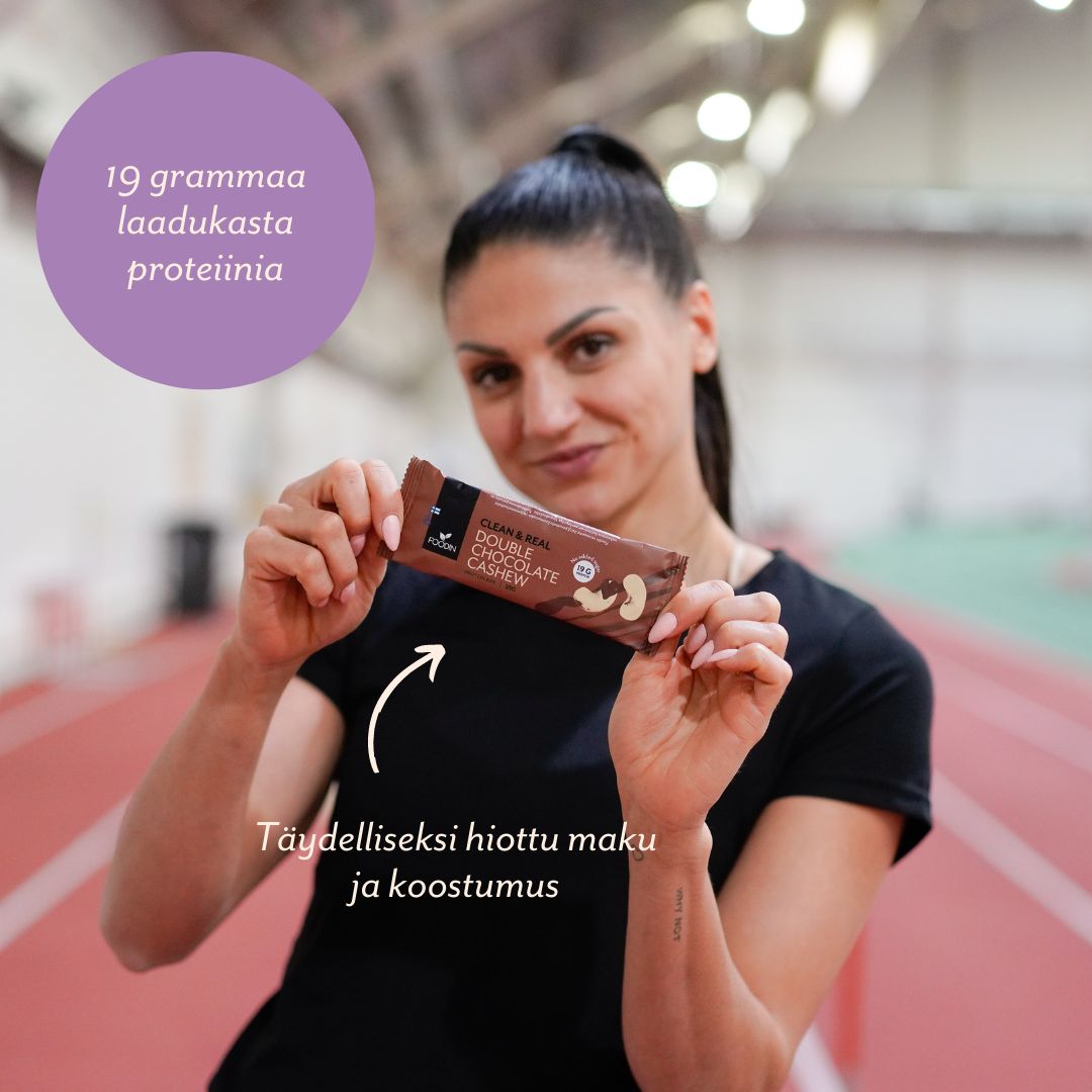 A woman in sportswear holds a rectangular protein bar in shiny, branded packaging at an indoor track. The packaging features clear, bold Finnish text: 19 grammaa laadukasta proteiinia and Täydelliseksi hiottu maku ja koostumus. Background shows athletic flooring. Shot is waist-up with focus on the hands and product.