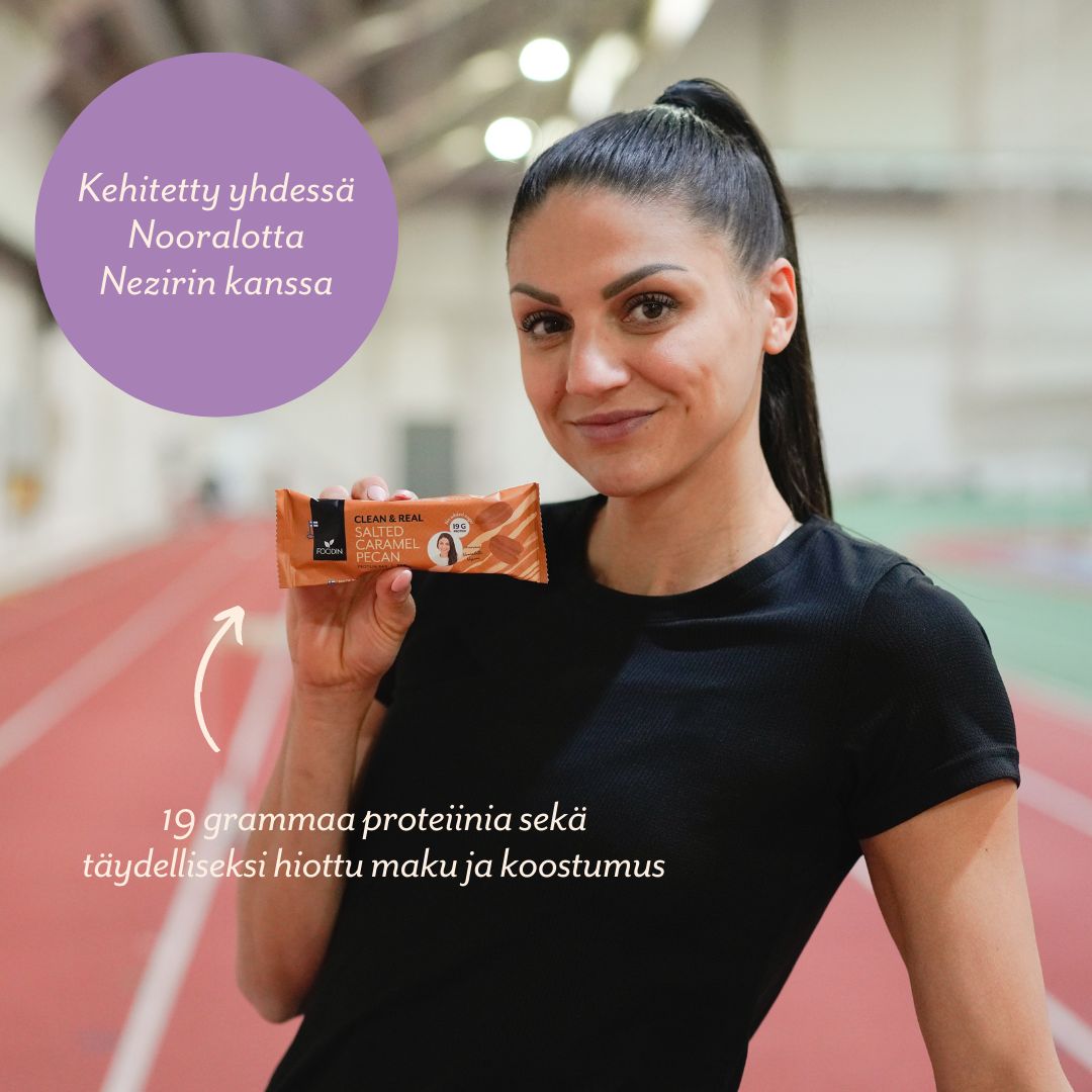 A woman in sportswear stands on an indoor running track, smiling and holding a protein bar. The bar in her hand is rectangular, in branded packaging with both matte and glossy areas. Legible Finnish text on the packaging describes high-protein content and mentions Nooralotta Neziri collaboration. A well-lit indoor sports facility is visible in the background.
