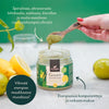 A hand scoops green powder from a jar labeled Green Superfood by Foodin. The jar is cylindrical with a black lid and matte-finish label. Lemons, leafy greens, and a metal spoon are placed nearby on a flat light-colored surface. Distinctive Finnish text in sans-serif font appears on the label. A hand is present interacting with the product.
