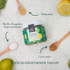 A jar of Foodin Green Superfood is surrounded by lemons, a lime, wooden spoons, and white powder. The jar is cylindrical with a matte black lid; the label features clear sans-serif text and green accents. Arrows point to labeled ingredients: yerba mate, lion’s mane, Bacillus Coagulans bc30 -probiotti. Text reads: BOOSTIA SEKOITUKSEEN TUOVAT.. The background is plain white. No people or hands are present. Shot is from above.