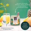 A jar of Foodin Green Superfood powder sits on a table with a glass of green juice, surrounded by fruits and fresh ingredients. The plastic jar is cylindrical with a matte label featuring sans-serif Finnish text summarizing key plant-based ingredients. The scene is well-lit with the background softly blurred. No people or hands are present in the image.