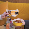 Foodin Honey Garlic Almonds; kaksi kättä pitelee mattapintaista muovipussia, jossa lukee ”honey garlic almonds” lihavoidulla groteskilla, sekä pyöreää keltaista keraamista kulhoa täynnä samaa naksua; kirkas muovinen juomapullo valkoisella korkilla ja toinen suljettu naksupussi vaalealla puisella suorakaiteen muotoisella pöydällä; taustalla verhoiltu kirkkaankeltainen sohva; kuva ylhäältä, ei kasvoja näkyvissä.