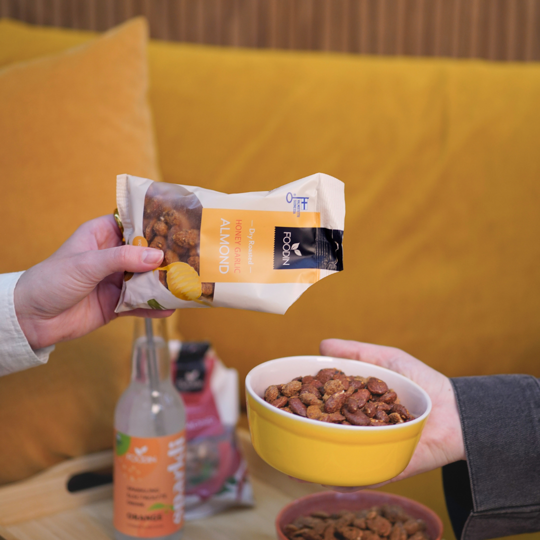 Foodin Honey Garlic Almonds; kaksi kättä pitelee mattapintaista muovipussia, jossa lukee ”honey garlic almonds” lihavoidulla groteskilla, sekä pyöreää keltaista keraamista kulhoa täynnä samaa naksua; kirkas muovinen juomapullo valkoisella korkilla ja toinen suljettu naksupussi vaalealla puisella suorakaiteen muotoisella pöydällä; taustalla verhoiltu kirkkaankeltainen sohva; kuva ylhäältä, ei kasvoja näkyvissä.