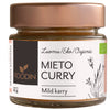 A glass jar of Foodin brand organic mild curry spice blend, 25g, with a copper-colored metallic screw-on lid and a cylindrical shape. The white matte label wraps around the jar, displaying botanical illustrations and bilingual Finnish and English text in simple sans-serif fonts. The product stands on a plain surface with no visible background elements.