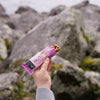 A hand holds a rectangular nut bar sealed in a glossy purple plastic wrapper. The wrapper features bold, white sans-serif lettering and partial product information. The hand is centered in the frame with visible fingers grasping the bar. In the background, there are large rocks covered in green moss in an outdoor setting. A hand holding a nut bar with a purple wrapper stands out against large, moss-covered rocks outdoors, highlighting the snack’s appeal in nature.