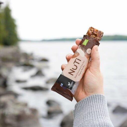 A hand holding a NuTi chocolate chip nut bar with a blurred lakeside and rocky, nature-filled shore in the background. The bar is packaged in a glossy wrapper showing the product name “NuTi” and details about chocolate chips and nuts. The shape is rectangular with visible edges through the packaging. Block-style typography with contrasting colors is used on the label.