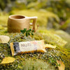 A wooden cup with a smooth finish and round opening sits beside a rectangular, glossy, plastic-wrapped NuTi nut bar. The nut bar packaging is clear with white and brown block text displaying “NuTi.” Both items rest on dense green moss with scattered small leaves and pieces of twigs; outdoor scene. A wooden cup and a packaged NuTi nut bar rest on green moss, surrounded by small leaves and earthy outdoor elements.