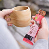 A person holds a wooden cup of coffee in one hand and an unwrapped NUTI organic berry snack bar in the other, set against a scenic outdoor background. The snack bar packaging is white with green and red accents, matte finish, and rectangular shape; only “NUTI organic berry” is clearly legible. The cup has a smooth texture and natural wood grain. Medium close-up shot.