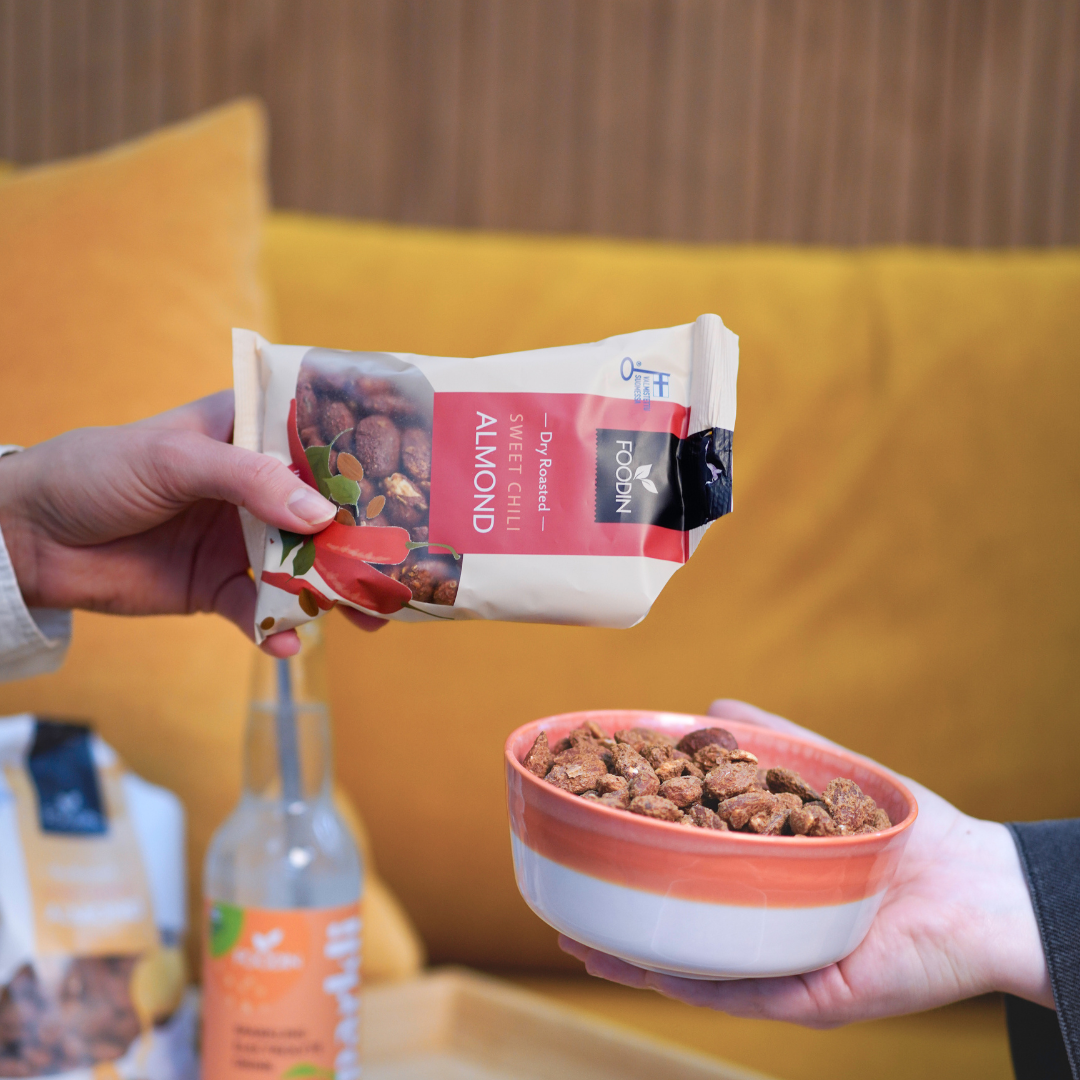 Foodin Sweet Chili Almonds; henkilö ojentaa mattapintaisen, suorakaiteen muotoisen muovipussin, jossa lukee ”dry roasted sweet chili almonds” lihavoidulla groteskilla, toiselle henkilölle, joka pitelee pyöreää valkoista kulhoa täynnä manteleita; taustalla näkyvissä pullo ja tekstiilityynyjä; kuva edestä, molemmat kädet näkyvissä.