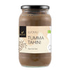 Foodin Luomu Tahini Tumma -lasipurkki; musta muovinen kansi; sylinterimäinen purkki, jota kiertää mattapintainen beige-ruskea etiketti tekstillä "Luomu Tumma Tahini" selkeällä tekstausvapaalla kirjasimella; kuvattu silmän korkeudelta yksiväristä taustaa vasten.