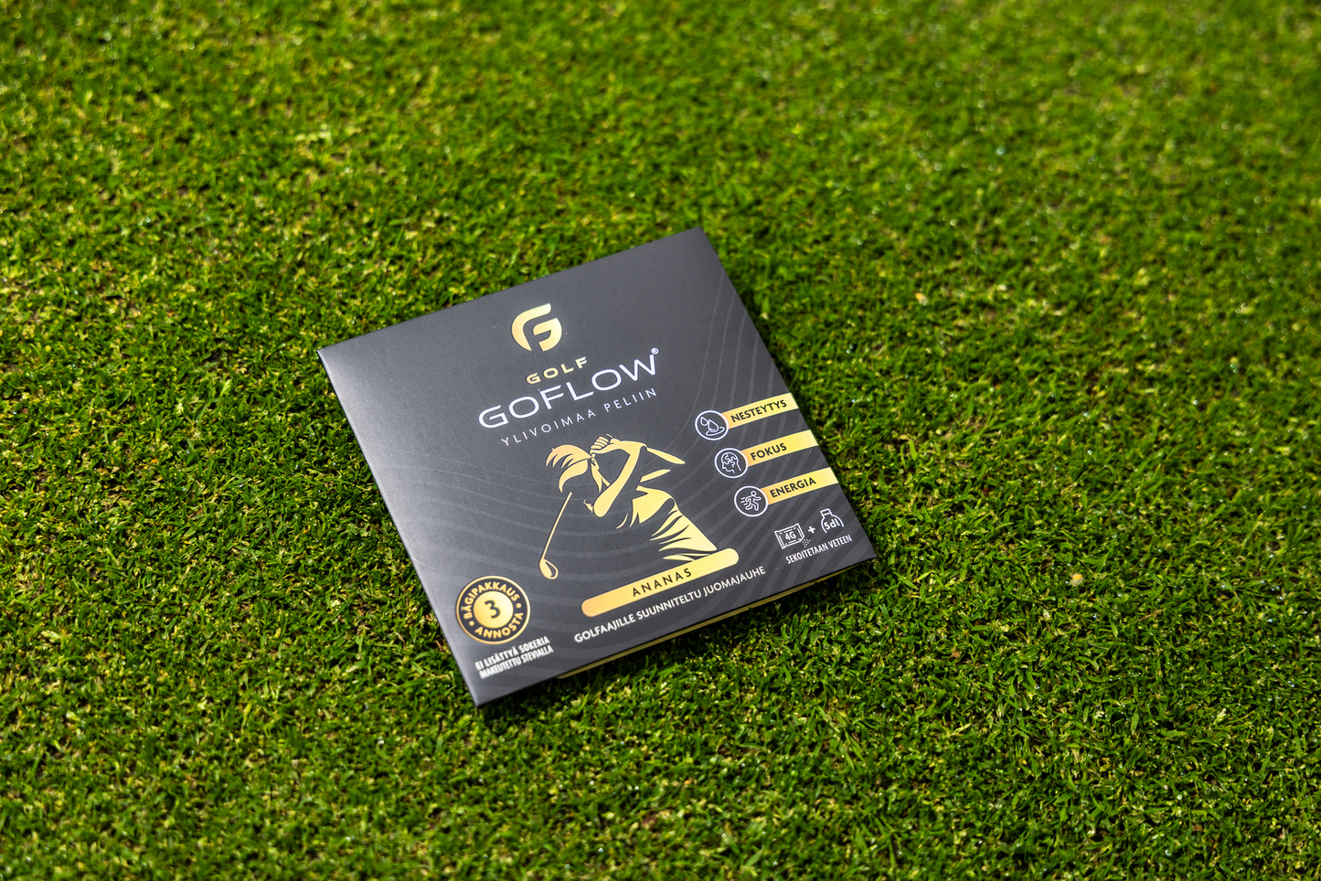 Golf Goflow Mini; musta ja kultainen GoFlow-golfvälinepakkaus vihreällä nurmikolla; suorakulmainen rasia mattapinnalla näyttää valkoisen golfaajan siluetin, useita logoja, palkkimaista tuotetietoa ja tekstin ”ANANAS” lihavoiduin suuraakkosin keltaisella raidalla lähellä alareunaa; ei ihmisiä tai käsiä kuvassa; kuva otettu hieman korotetusta kulmasta.