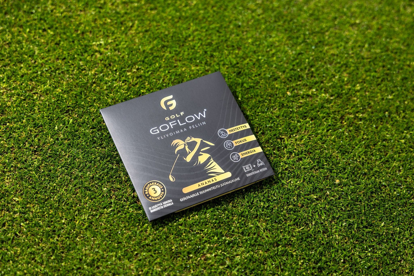 Golf Goflow Mini; musta ja kultainen GoFlow-golfvälinepakkaus vihreällä nurmikolla; suorakulmainen rasia mattapinnalla näyttää valkoisen golfaajan siluetin, useita logoja, palkkimaista tuotetietoa ja tekstin ”ANANAS” lihavoiduin suuraakkosin keltaisella raidalla lähellä alareunaa; ei ihmisiä tai käsiä kuvassa; kuva otettu hieman korotetusta kulmasta.