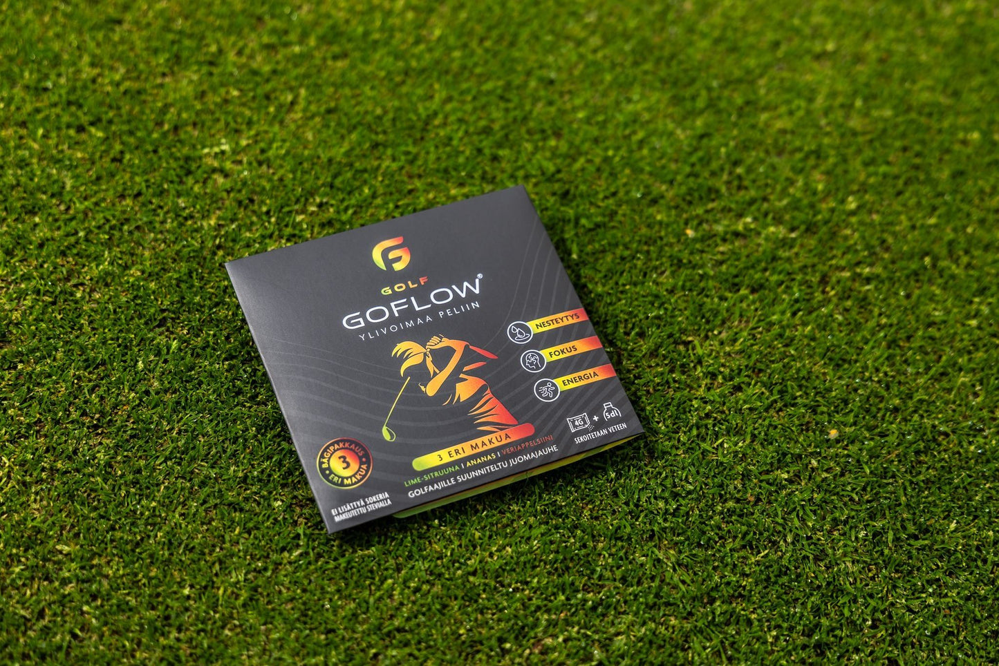 Golf Goflow Mini; musta suorakulmainen rasia tekstillä ”GOFLOW golf strings” seisoo kirkkaan vihreällä tekonurmella; kiiltävässä pakkauksessa monivärinen golfaajakuvitus ja tuotetietoa useilla kielillä; kirjasin sisältää lihavoitua groteskia ja kursiivia; ei ihmisiä, käsiä tai toimintaa kuvassa.