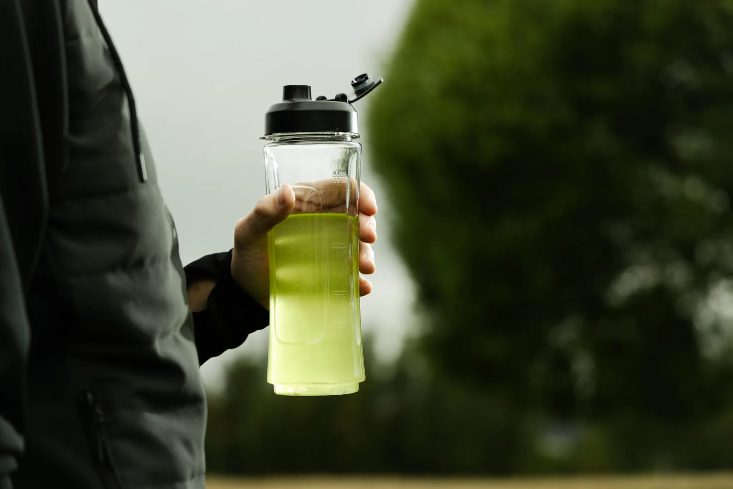Golf Goflow Mini; henkilö tummassa takissa pitelee kirkasta sylinterin muotoista lasipulloa täynnä kirkkaanvihreää nestettä; pullossa ei näy etikettiä tai korkin yksityiskohtia; taustalla epätarkkoja lehtevä puustoa ja vihreyttä; ei luettavaa tekstiä näkyvissä; kuva lähikuvana näyttäen vain kädet ja osan vartaloa.