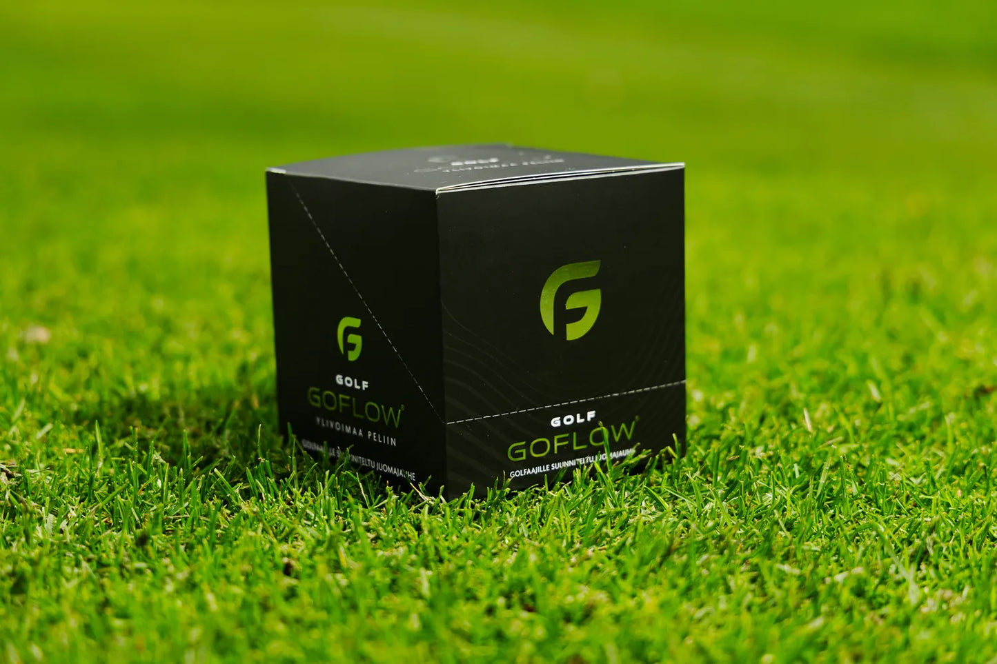 Golf Goflow Pro; suorakulmainen musta pahvirasia mattapinnalla ja suuri vihreä G-logo etupuolella; lisäksi vihreä ”GOLF GOFLOW” -brändäys lihavoidulla groteskikirjasimella; rasia sinetöity terävin reunoin ilman näkyviä avauksia tai lisätarvikkeita; kirkas tekonurmitausta muistuttaa golfkenttää ilman ihmisiä tai esineitä; kuva otettu suoraan edestä tasaisessa valaistuksessa.