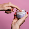 Goliate 2-in-1 Makuliukuvoide Vanilja; pakkauskuva, jossa henkilö kastaa sormen kirkkaaseen pyöreään muovipurkkiin valkoisella kierrekannella; purkin etiketissä lukee ”Couple Gourmand Lubricating Cream” yksinkertaisella mustalla groteskitekstillä; astia lepää tasaisella vaaleanpunaisella pinnalla, kuvattu läheltä; tausta yhtenäisen vaaleanpunainen.