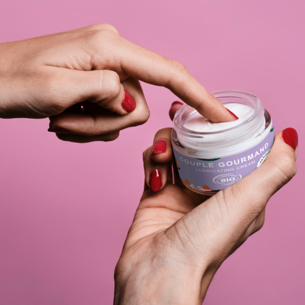 Goliate 2-in-1 Makuliukuvoide Vanilja; pakkauskuva, jossa henkilö kastaa sormen kirkkaaseen pyöreään muovipurkkiin valkoisella kierrekannella; purkin etiketissä lukee ”Couple Gourmand Lubricating Cream” yksinkertaisella mustalla groteskitekstillä; astia lepää tasaisella vaaleanpunaisella pinnalla, kuvattu läheltä; tausta yhtenäisen vaaleanpunainen.