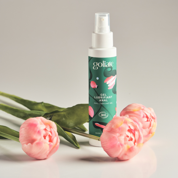 Goliate Anaaliliukuvoide; pullo Goliath-liukuvoidegeeliä vihreällä etiketillä seisoo pystyssä kolmen vaaleanpunaisen tulppaanin vieressä sileällä, vaaleasävyisellä pinnalla; pullo sylinterin muotoinen läpikuultavaa tai läpinäkyvää materiaalia mattapinnalla ja valkoisella korkilla; vihreässä etiketissä yksinkertaista mustaa ja valkoista groteskitekstiä; tausta neutraali, beige tai harmaa sävy; ei ihmisiä tai käsiä kuvassa.