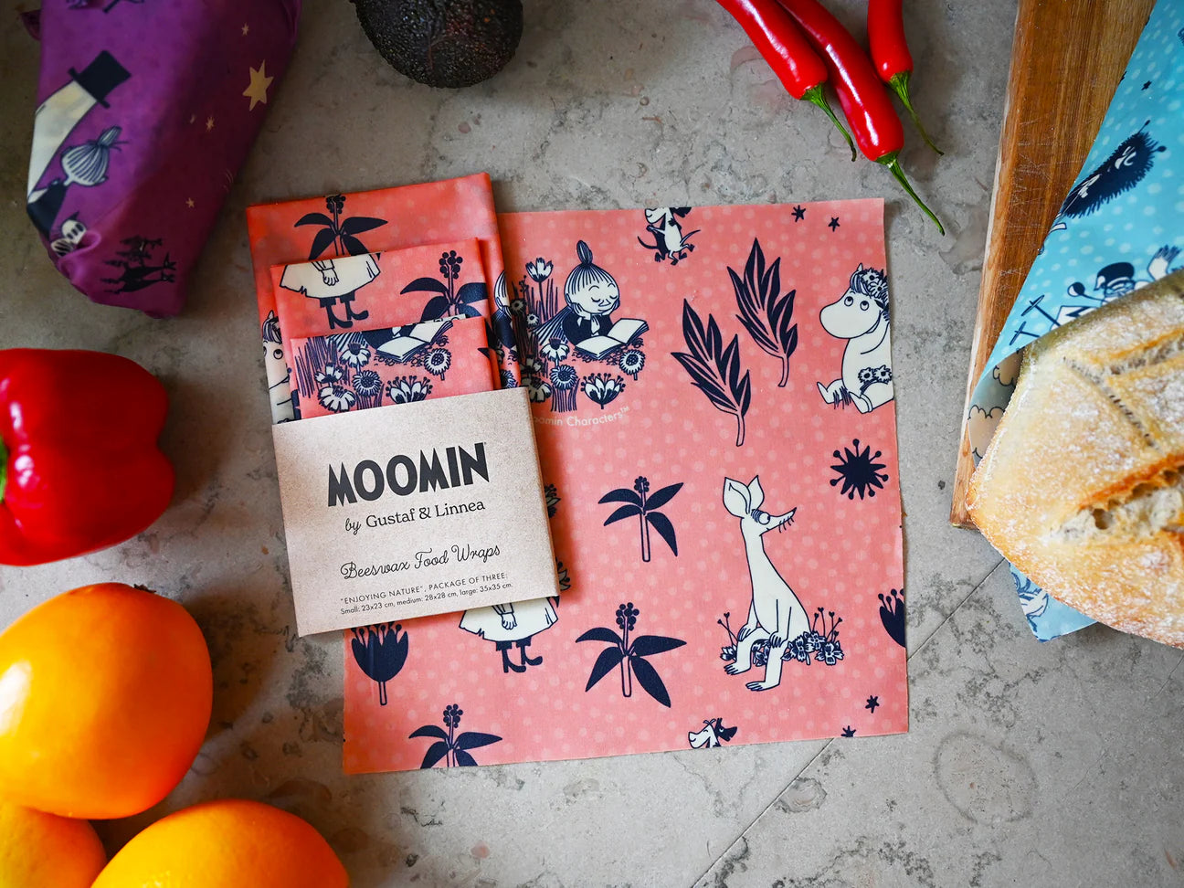 Gustaf & Linnea Muumi Mehiläisvahakääre 3-pack; Muumi-hahmoin koristeltu mehiläisvahakääre lepää keittiön tasolla ympärillään leipää, punaista paprikaa, appelsiineja, avokadoa ja chilipapruikoita; vieressä taitettu yhteensopiva kääre, merkitty MOOMIN by Gustaf & Linnea; kääreet ovat suorakulmaisia värikkäin painetuin kuvioin ja mattapintaisia; etiketissä yksinkertainen groteskifontti; ympäristönä keittiön taso ilman ihmisiä tai käsiä kuvassa.