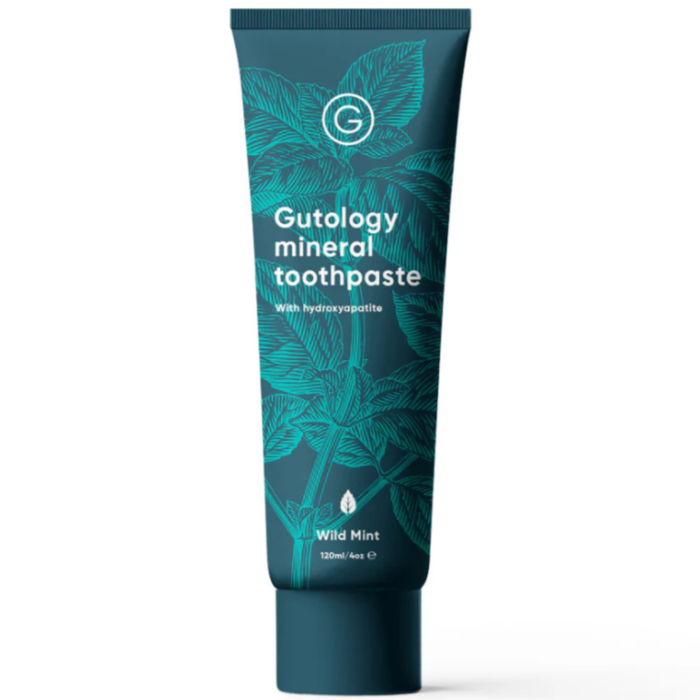 Gutology Hammastahna; tuubi Gutology-mineraalihammastahnaa villiminttumaussa, kasviaiheisin lehtikuvituksin tummanvihreällä taustalla; 120 ml:n (4 oz) tuubi korostaa hydroksiapatiittia ja mineraaleja; tuubi on sylinterinmuotoinen valkoisella korkilla ja mattapinnalla; groteskikirjasin etiketin tekstissä; ei ihmisiä tai käsiä näkyvissä; kuva keskittyy suoraan tuotteeseen yksivärista taustaa vasten.