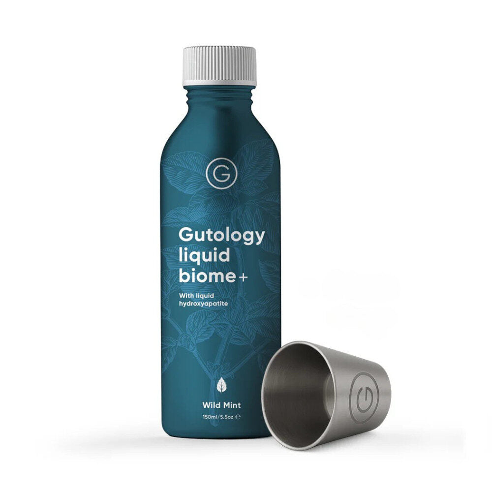 Gutology Liquid Biome+ Suuhuuhde; sininen pullo tekstillä Gutology liquid biome+ Wild Mint pystyssä ruostumattomasta teräksestä valmistetun, kyljellään olevan mukin vierellä valkoisella taustalla; pullo sylinterin muotoinen valkoisella kierrekorkilla, etiketissä valkoista ja vihreää groteskitekstiiä; teräsmuki hartsattu pinta; ei ihmisiä tai toimintaa kuvassa.