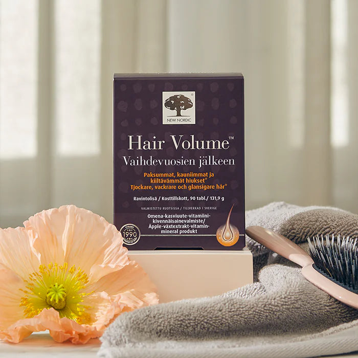 Hair Volume Vaihdevuosien jälkeen; laatikko ravintolisää pöydällä persikankukan, harmaan pyyhkeen ja hiusharjan vieressä pehmeästi valaistussa ympäristössä; laatikko on mattapintainen syvän violetti väriltään, valkoista tekstiä selkeällä groteskikirjasimella; etiketissä lukee tuotenimi; pakkaus on suorakulmainen; ei ihmisiä tai käsiä näkyvissä.