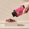 Hair, Skin & Nail Strong Gummies; käsi kaataa purutabletteja vaaleanpunaisella etiketillä varustetusta muovipullosta pyöreään lasiastiaan, jossa punaisia karhunmuotoisia purutabletteja; astia lepää mattabeigellä suorakaiteen muotoisella alustalla; etiketissä pääosin valkoista groteskitekstiä ja pienempää tekstiä; tausta vaalea ja yksinkertainen; ei muuta rekvisiittaa tuotteen, astian ja alustan lisäksi; lähikuva.