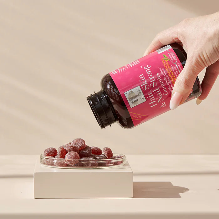 Hair, Skin & Nail Strong Gummies; käsi kaataa purutabletteja vaaleanpunaisella etiketillä varustetusta muovipullosta pyöreään lasiastiaan, jossa punaisia karhunmuotoisia purutabletteja; astia lepää mattabeigellä suorakaiteen muotoisella alustalla; etiketissä pääosin valkoista groteskitekstiä ja pienempää tekstiä; tausta vaalea ja yksinkertainen; ei muuta rekvisiittaa tuotteen, astian ja alustan lisäksi; lähikuva.