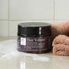Hair Volume Repair Mask; musta sylinterinmuotoinen purkki valkoisin groteskikirjaimin on aseteltuna kiiltävälle, märälle kylpyhuonepinnalle osittain saippuavaahdon peitossa; vieressä taitettu persikanvärinen pyyhe ja pieni asetelma näkyviä ravitsevia ainesosia; tausta koostuu kiiltävistä valkoisista suorakulmaisista laatoista; ei ihmisiä tai käsiä näkyvissä.