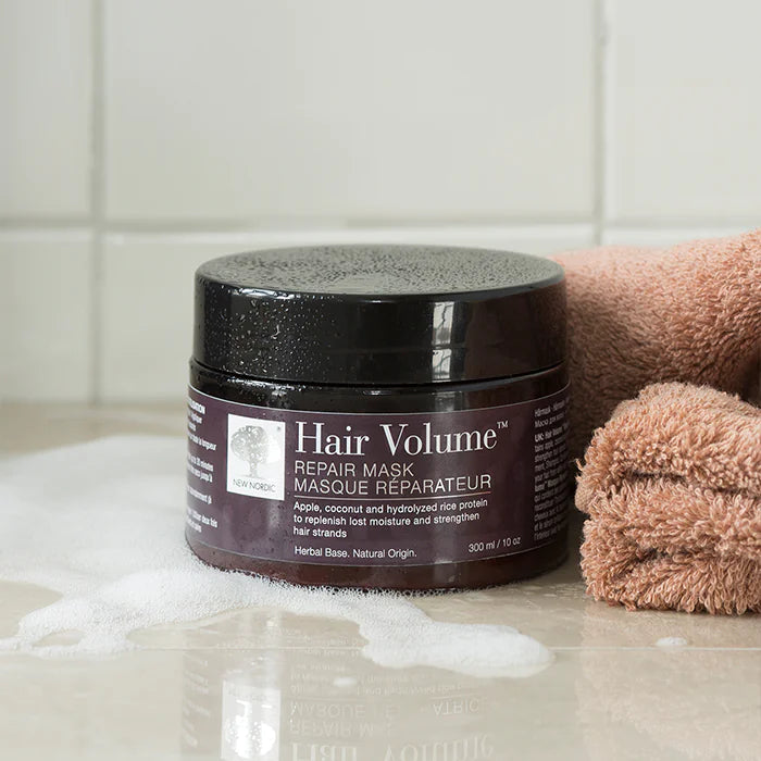 Hair Volume Repair Mask; musta sylinterinmuotoinen purkki valkoisin groteskikirjaimin on aseteltuna kiiltävälle, märälle kylpyhuonepinnalle osittain saippuavaahdon peitossa; vieressä taitettu persikanvärinen pyyhe ja pieni asetelma näkyviä ravitsevia ainesosia; tausta koostuu kiiltävistä valkoisista suorakulmaisista laatoista; ei ihmisiä tai käsiä näkyvissä.