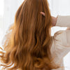 Hair Volume Vaihdevuosien jälkeen; henkilö, jolla on pitkät aaltoilevat punaiset hiukset, vetää sormiaan hiuksissaan; hänellä on yllään kermanvärinen, pitkähihainen yläosa; kuva on otettu takaapäin ja rajattu näyttämään yläselkä ja pää; tausta on pehmeästi valaistu lämpimin sävyin; kuvassa ei näy pintoja, etikettejä, pakkausta tai brändimerkintöjä.