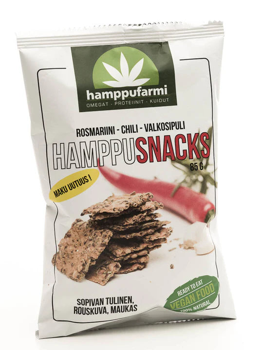 Hamppufarmi HamppuSnacks Rosmariini-Chili-Valkosipuli; pussi hamppuvälipaloja rosmariini-, chili- ja valkosipulimauilla; suorakulmainen mattapintainen pussi vihreä-valkoisena, etuosassa kuvia pyöreistä välipaloista ja kokonaisesta punaisesta chilistä; vegaaniset ruokamerkit pyöreinä kuvakkeina; lihavoitu groteskiteksti sisältää tuotenimen ja maun; tausta yksivärinen, ei ihmisiä tai käsiä näkyvissä, kuva otettu edestäpäin studiossa.