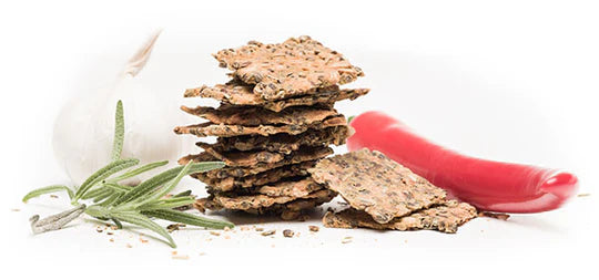 Hamppufarmi HamppuSnacks Rosmariini-Chili-Valkosipuli; pino suorakulmaisia ruskeita siemenrapeleipiä mattapinnalla valkoisella taustalla; leipien vieressä kokonainen valkosipulinkynsi, tuore punainen chili ja rosmariininoksa; ei pakkausta tai typografiaa näkyvissä; ei ihmisiä tai toimintaa kuvassa.