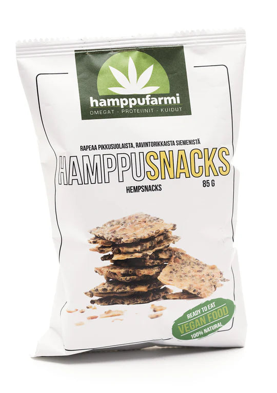 Hamppufarmi HamppuSnacks; valkoinen pussi vegaanisia hampunsiemensnackseja, näyttää pinottuja näkkileipäpaloja; pakkauksessa vihreää ja keltaista tekstiä sekä lehtilogo, maininnat valmiiksi syötävästä, vegaanisesta, gluteenittomasta ja 100-prosenttisesta luonnollisuudesta; pinta mattainen ilman kiiltoa; typografia lihavoitua ja groteskia; ei ihmisiä tai käsiä; vaalea tausta, tiiviisti rajattu tuotekuva.