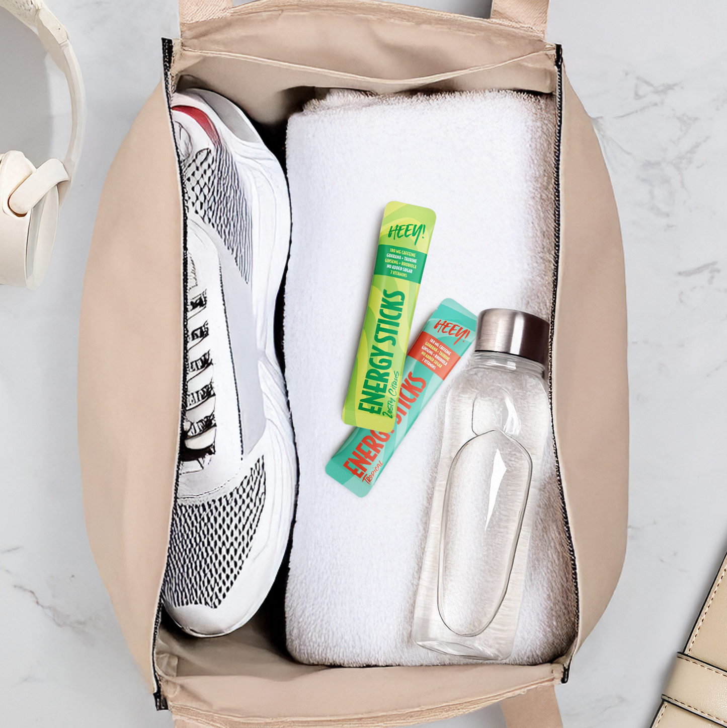 Heey! Energy Sticks Caffeine + Guarana, maku Tropical; avoin kuntosalilaukku valkoisen pyyhkeen, valkoisten lenkkitossujen, kahden energiapatukkapussin ja läpinäkyvän uudelleentäytettävän vesipullon kanssa, aseteltuna vaalealle alustalle; laukku kangasta vetoketjuavauksella; tossut nauhalliset matalavartiset; vesipullo lieriömäinen läpinäkyvä muovi kierrekannella; energiapussit suorakaiteen muotoisin kääntein, vähäistä näkyvää tekstiä; ei ihmisiä tai käsiä.
