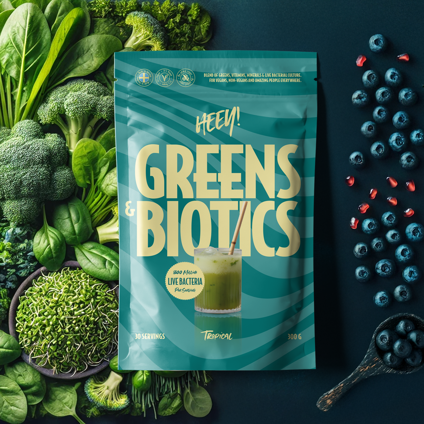 Heey! Greens & Biotics Superfood Mixed Berries: matta turkoosi pussi tekstillä "Greens & Biotics" tummalla, sileällä alustalla; pussissa lohko- ja pyöristetty kirjasin sekä piirroskuva lasista vihreää nestettä pillillä; edessä ja ympärillä tuoretta parsakaalia, pinaatinlehtiä, lehtikaalia, ituja ja mustikoita; ei ihmisiä tai toimintaa näkyvissä.