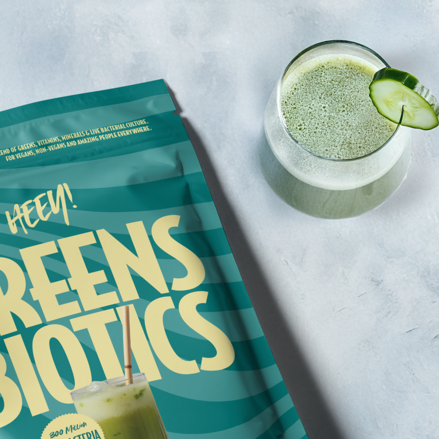 Heey! Greens & Biotics Superfood Mixed Berries: lasi vihreää smoothieta kurkkuviipaleella reunalla mattapintaisen pussin vieressä tekstillä "Greens Biotics powder"; pussi on suorakulmainen ja pääosin vaaleansävyinen groteskikirjasimella; molemmat esineet lepäävät vaalealla, kevyesti tekstuurisella pinnalla; ei ihmisiä tai käsiä näkyvissä, kuva otettu ylhäältä.