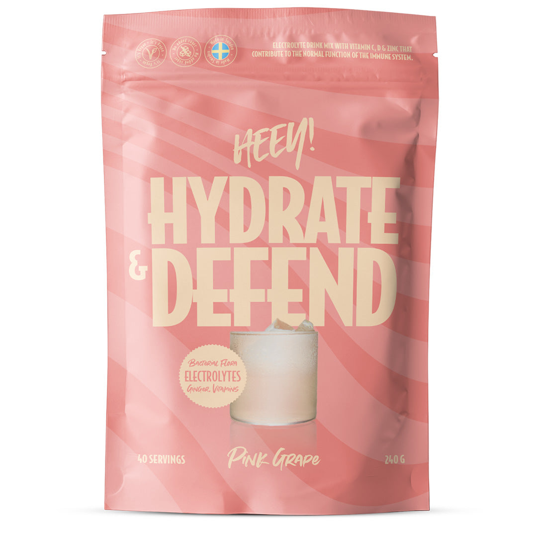 Heey! Hydrate & Defend Electrolytes + Immune Boost; vaaleanpunainen, mattapintainen uudelleensuljettava pussi, jossa lukee "Hydrate & Defend", etupuolella kuvitus lasista pinkkiä rypälejuomaa; pakkauksessa mainitaan elektrolyytit, vastustuskyvyn tuki, 40 annosta ja 240 g; teksti groteskifontilla eri lihavuuksin ja koin; pussi seisoo pystyssä, ei ihmisiä tai käsiä näkyvissä yksivärisessä studiokuvassa.