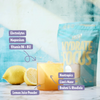 Heey! Hydrate & Focus Electrolytes, maku Lemon; lasillinen keltaista juomaa sitruunan kanssa, etiketöity ainesosin kuten elektrolyytit, vitamiinit ja nootropit, etualalla, taustalla Hydrate & Focus -juomajauhepussi; molemmissa vaaleanvioletit etiketit erityisominaisuuksin; pakkaus yksittäisannos, pussissa modernia groteskitekstiä; ei ihmisiä tai käsiä; siisti studioasetelma, kuvattu suoraan edestä.