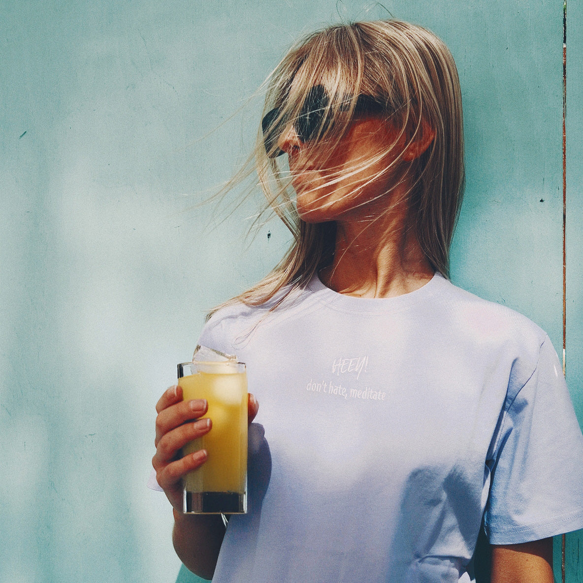 Heey! Hydrate & Focus Electrolytes Raspberry Lemonade; pitkähiuksinen vaaleahiuksinen henkilö tummissa aurinkolaseissa ja laventelinvärisessä puuvillaisessa tai vastaavassa collegepaidassa, jossa lukee pienaakkosin pyöristetyllä groteskilla "don't hate, meditate"; henkilö pitelee kirkasta lasia, jossa appelsiinimehua; hiussuortuvia tuulee kasvojen editse; tausta yksivärinen vaaleansininen.