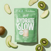 Vihreä Hydrate & Glow -juomajauhepussi on aseteltu vaalealle pinnalle, ympärillä viipaloituja vihreitä omenoita ja kiivejä. Pussi on mattapintainen, suorakulmainen ja tiivistetyllä yläreunalla. Lihavoitu valkoinen pääteviivaton typografia näyttää tekstin Hydrate & Glow ja mainitsee elektrolyytit nesteytyksen tueksi sekä maun green apple & kiwi. Ihmisiä tai käsiä ei näy kuvassa. Kuva on otettu ylhäältä.