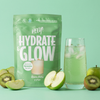 Vihreä Hydrate & Glow -pussi on aseteltu vihreän juoman ja jään täyttämän lasin viereen, ympärillä viipaloituja vihreitä omenoita ja kiivejä mintunvihreällä taustalla. Pussi on mattapintainen ja siinä on yksinkertaista valkoista pääteviivatonta tekstiä. Kuva on otettu ylhäältä, ja sisältö on aseteltu siististi ilman ihmisiä tai käsiä.