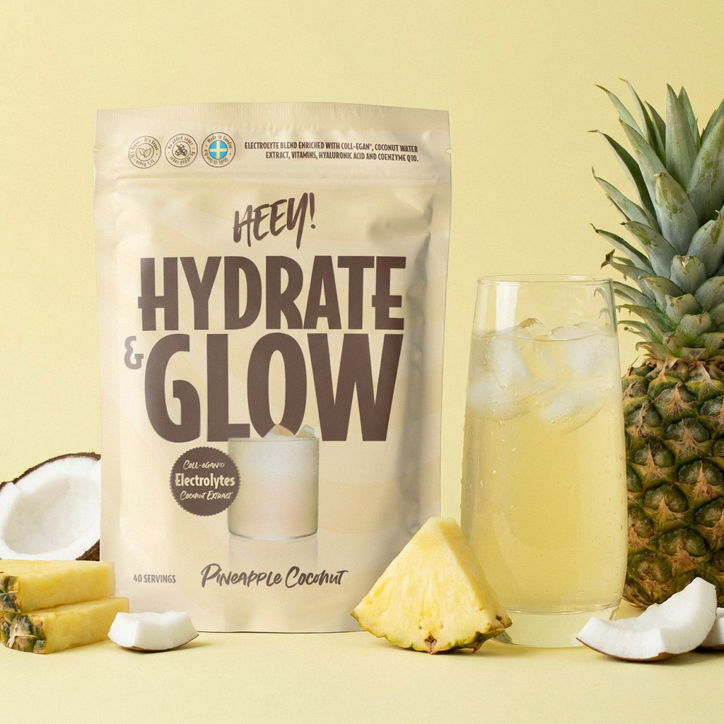 Suorakulmainen, mattapintainen pussi näyttää tekstin Hydrate & Glow selkeällä pääteviivattomalla fontilla. Sen vieressä on läpinäkyvä lasi täynnä vaaleankeltaista nestettä. Vierellä on aseteltu ananasviipaleita, kookospaloja ja kokonainen ananas tasaiselle pinnalle vaalean keltaista taustaa vasten. Ihmisiä tai käsiä ei näy kuvassa.