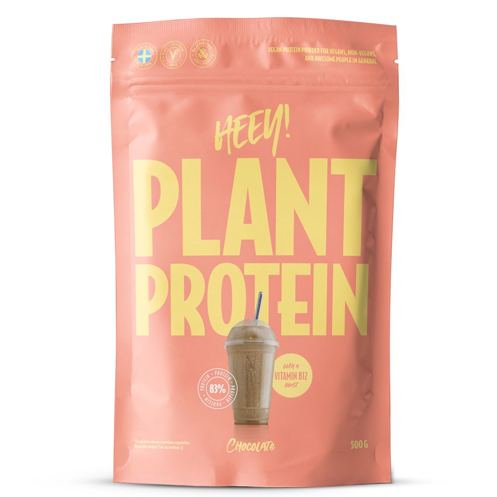 Heey! Plant Protein, maku Chocolate; koralliväristä pinkki pussi tekstillä HEY! PLANT PROTEIN isoin keltaisin kirjaimin, kuva suklaanmakuisesta smoothiesta, esillä B12-vitamiini ja 83 % kasviproteiinipitoisuus; pakkauskoko 500 g; pinta mattapintainen; joustava muovipakkaus lämpösaumatulla yläreunalla; teksti lihavoitua groteskia, keskitettynä etuosaan; valkoinen tausta, ei ihmisiä tai esineitä.