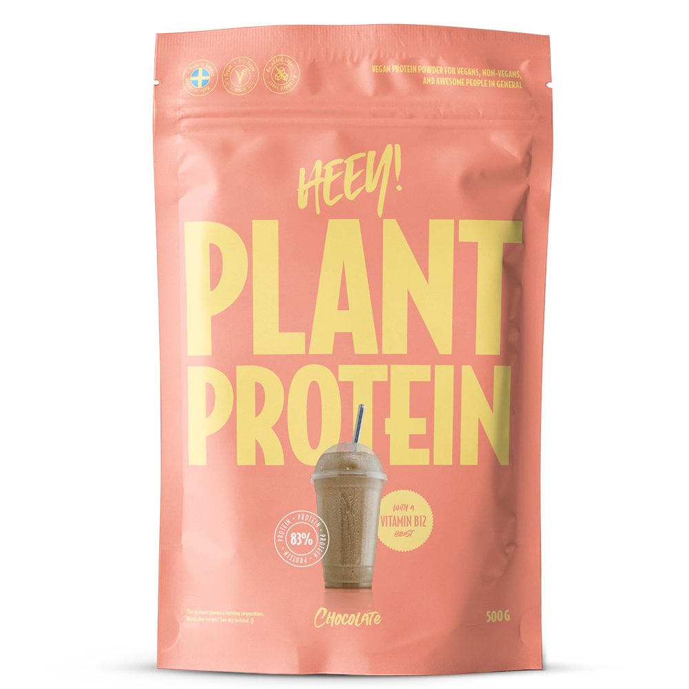 Heey! Plant Protein, maku Chocolate; koralliväristä pinkki pussi tekstillä HEY! PLANT PROTEIN isoin keltaisin kirjaimin, kuva suklaanmakuisesta smoothiesta, esillä B12-vitamiini ja 83 % kasviproteiinipitoisuus; pakkauskoko 500 g; pinta mattapintainen; joustava muovipakkaus lämpösaumatulla yläreunalla; teksti lihavoitua groteskia, keskitettynä etuosaan; valkoinen tausta, ei ihmisiä tai esineitä.