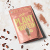 Heey! Plant Protein, maku Chocolate; pussi nimellä Plant Protein keltaisin tekstein lepää marmoripinnalla, ympärillä kokonaisia kaakaopapuja, irtokaakaojauhetta ja pakkaukseen painettu kuva suklaisesta proteiinishakesta; pussi suorakaiteen muotoinen mattapinnalla; typografia lihavoitu ja groteski; ei ihmisiä tai käsiä; yläviistosta kuvattu neutraalissa valaistuksessa.