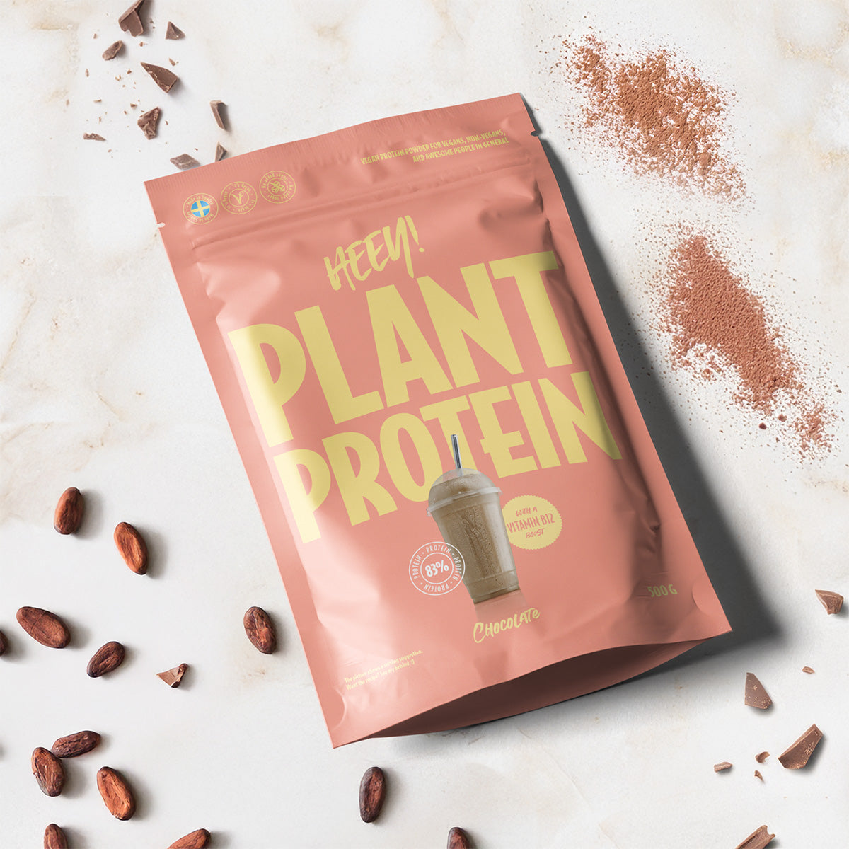 Heey! Plant Protein, maku Chocolate; pussi nimellä Plant Protein keltaisin tekstein lepää marmoripinnalla, ympärillä kokonaisia kaakaopapuja, irtokaakaojauhetta ja pakkaukseen painettu kuva suklaisesta proteiinishakesta; pussi suorakaiteen muotoinen mattapinnalla; typografia lihavoitu ja groteski; ei ihmisiä tai käsiä; yläviistosta kuvattu neutraalissa valaistuksessa.