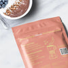 Heey! Plant Protein, maku Chocolate; suklaasmoothiekulho granolan ja kookoshiutaleiden kera proteiinijauhepussin vieressä, jossa esitellään herkullisia smoothie- ja shake-reseptejä; lusikka ja muutama mustikka vieressä; smoothie tummanruskeaa pyöreässä valkoisessa kulhossa, pinnalla siististi aseteltu granolaa ja valkoisia kookoshiutaleita