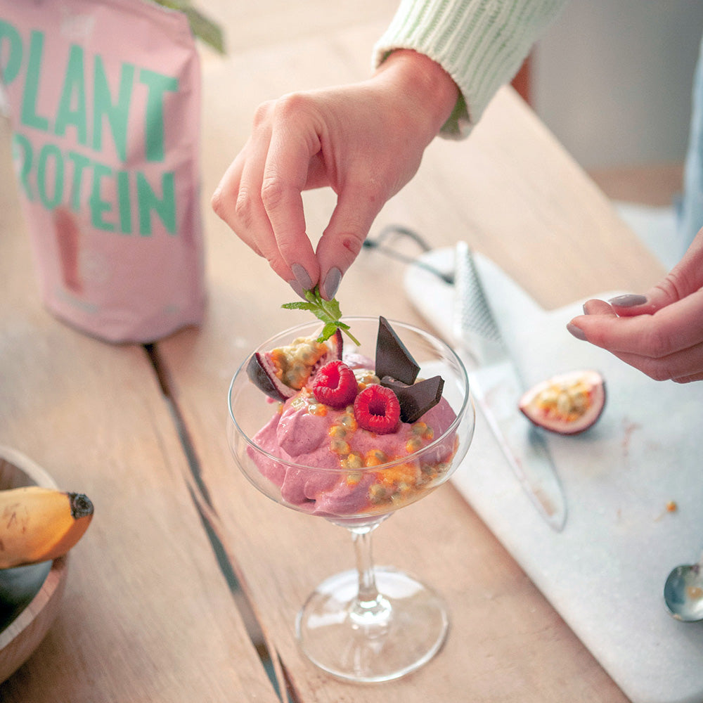 Heey! Plant Protein, maku Strawberry; henkilö koristelee vaaleanpunaista smoothiekulhoa lasissa mintulla, vadelmilla ja suklaanpaloilla; läpinäkyvä lusikka näkyvissä; kasviproteiinipussi seisoo pystyssä puupöydällä, mattapintainen pakkaus yksinkertaisin painetuin tekstein, osittain luettavissa Protein; leikattua hedelmää aseteltuna kulhon viereen; neutraali sisätila ilman muita näkyviä taustaelementtejä.