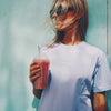 Heey! Plant Protein, maku Strawberry; tuote on kylmä vaaleanpunainen smoothie kirkkaassa muovimukissa litteine kansineen ja pillillä; sitä pitelevällä naisella tuulessa liehuvat vaaleat hiukset ja aurinkolasit; hänellä vaaleanvioletti mattapintainen puuvillapaita mustin lihavoiduin tekstein: HEEY! don't hate, meditate; tausta sileä yhtenäinen sininen seinä; kuva vyötäröstä ylöspäin ulkona.